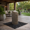 A stone pillar fountain with a small white spout sits on a bed of dark river stones on a stone patio, with a covered porch, outdoor sofa, and landscaped garden in the background.