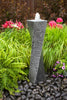 A modern twisted stone waterfall fountain with a textured dark gray body and a white water spout, set in a garden on a bed of dark river stones surrounded by green ferns, ornamental grass, and pink flowers.