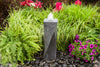A tall gray stone fountain with a textured surface and a small white water spout, set in a garden bed surrounded by green ornamental grass, ferns, and pink flowers on dark river rocks.
