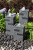 Three rectangular stone water fountains with textured gray surfaces and clear water spouts, arranged on dark river rocks surrounded by green ferns and purple flowers in a garden setting.