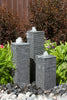 Three-tiered gray stone fountain garden statues with flowing water, set in a landscaped garden bed with green ferns, small flowers, and dark river rocks.