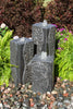 Three dark gray stone waterfall fountains arranged in a garden setting, surrounded by green ferns, colorful flowers, and small multicolored pebbles.