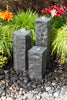 Three dark gray stone fountain posts with small water fountains at the top, set in a garden bed on dark river stones with green ornamental grass and colorful flowers in the background.
