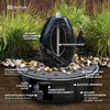 Black stone fountain with a central oval opening and visible silver tubing, shown outdoors on a pedestal base with labeled callouts about the fountain, pump, basin, and stones. The scene includes grass, pebbles, and a brown ground surface.