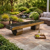 A rustic outdoor stone bench with a dark polished top and natural textured stone legs, placed on a paved patio in a landscaped garden setting with a small waterfall, plants, and a wicker sofa nearby.