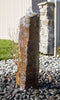 A natural brown stone waterfall fountain with a rough, polished block shape, set in a garden bed surrounded by black river rocks, green foliage, and colorful plants.