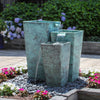 Three-tier rectangular outdoor water fountain with a textured turquoise-green patina finish, set in a garden bed with gray river stones and surrounded by flowers and green foliage.