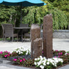 Three tall faux basalt stone fountain spires arranged in a cluster, shown outdoors in a garden setting with flowers, a stone wall, and patio furniture. A text banner at the top reads 'Extra Tall (24'-36'-48") Triple Faux "Basalt" Fountain Kit - LA7475'.
