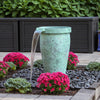 A freestanding cylindrical garden fountain with a textured mint-green body and a clear water stream spouting from the top, placed outdoors among bright pink flowers, dark pebbles, and a planter box in the background.
