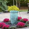 A freestanding cylindrical outdoor water feature with a textured turquoise-green finish and a curved spout from which water flows, set in a landscaped garden bed with bright pink flowers and dark gravel, on a stone patio with trees in the background.