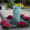 A freestanding cylindrical outdoor water feature with a flowing stream, finished in a textured turquoise-green patina. It is set in a landscaped garden bed with bright pink mums, gray river rocks, and a stone patio in the background.
