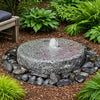 A round gray stone fountain with a small water jet, set in a garden bed surrounded by dark river rocks, mulch, and green plants.