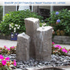 A short triple faux basalt fountain kit with three tall textured stone-like columns and flowing water, shown outdoors in a garden bed with dark river rocks, pink flowers, green foliage, and a house background. A blue text banner at the top reads 'Short (18"-24"-30") Triple Faux "Basalt" Fountain Kit - LA7400'.