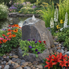 A natural stone fountain with a small waterfall spout, placed in a landscaped garden bed with colorful flowers, greenery, and a pond in the background.
