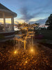 A decorative illuminated tree fountain or lighted garden sculpture with warm golden lights and hanging water-like strands, set in a pebble bed outdoors at dusk beside a gazebo and lawn.