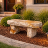 A rustic stone bench with a rectangular top and two thick block legs, placed in a landscaped garden bed with mulch, shrubs, ornamental grass, and a house exterior in the background.