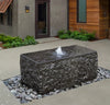 A rectangular black stone fountain with water cascading from the top, set in a landscaped garden bed with decorative rocks, in court yard entrance.