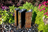 Three rectangular stone waterfall fountains with dark and rust-colored slabs, set in a garden on a bed of small pebbles, surrounded by green foliage and bright pink flowers.