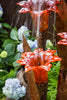 A close-up of a decorative garden fountain with translucent orange-red flower-shaped tiers and water flowing through them, set against green foliage and blue hydrangeas.