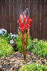 A modern garden fountain sculpture featuring six red calla lily flowers on brown metallic stems, set in a landscaped garden bed with pebbles, green plants, and a dark brown wooden fence background.