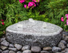 A round gray granite fountain stone with three small water jets, set in a garden bed surrounded by dark river rocks, green foliage, and bright pink flowers.