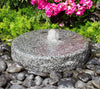 A round granite stone fountain with a small central water jet, set outdoors among dark river rocks, green foliage, and bright pink flowers.