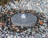 A round gray stone fountain or water feature with a small central stream, surrounded by small multicolored pebbles outdoors.