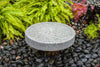 A round gray stone fountain basin with water flowing from the bottom, set in a garden bed surrounded by black river rocks, bright green ferns, and pink flowers.