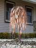 A decorative metal fountain sculpture shaped like a weeping tree, with copper-toned branches dripping water, installed outdoors in a bed of gray river rocks in front of a house with beige siding and dark-framed windows.