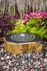 A round stone fountain with concentric ring patterns on top and a small central water jet, set in a garden bed with colorful flowers, green foliage, and pebbles.