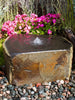 A rustic rectangular stone fountain basin with a small central water jet, set outdoors among pink flowers, green foliage, and small pebbles.