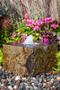 A rectangular stone fountain with a small central water jet, set outdoors on a bed of small multicolored pebbles with pink flowers and green foliage behind it.