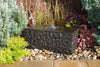 A rectangular black stone fountain with water cascading from the top, set in a landscaped garden bed with decorative rocks, green shrubs, and purple flowers against a dark wooden fence.