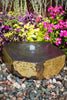 A hexagonal stone fountain basin with a small central water jet, set on a bed of multicolored river rocks in a garden bed surrounded by colorful pink flowers and green foliage.