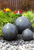 Three dark gray stone decorative spheres of different sizes arranged on a bed of small gray and white pebbles in a garden setting, with green grass, colorful flowers, and a brown fence in the background.