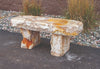 A rustic stone bench made from natural travertine or limestone, featuring a thick flat seat and two blocky legs, set outdoors on a paved surface with gravel and ornamental grass in the background.