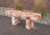 A rectangular stone bench made of orange and white marbled stone, set outdoors on a dark paved surface with gravel, ornamental grass, and a brown wall in the background.