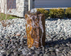 A rustic rectangular stone fountain with water flowing down its rough, textured surface, set outdoors on a bed of dark river rocks with gravel and shrubs in the background.