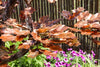 A copper leaf garden sculpture featuring realistic copper-colored oak leaves, shown outdoors with water spraying over it and purple flowers in the background against a dark wooden fence.