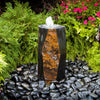 A natural stone fountain with a small waterfall spouting from the top, set in a bed of smooth river stones. The stone features a dual-tone design with a dark charcoal gray outer face and a warm, textured rust-orange and brown inner face.