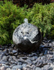 A polished black and white granite fountain sphere with a clear water jet spouting from the top, set outdoors on a bed of smooth river stones with green foliage in the background.