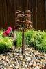 A decorative metal garden tree sculpture with layered brown and bronze leaves, planted in a stone bed with green grasses and bright pink flowers against a dark wooden fence.