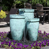 Three outdoor water fountains with a textured turquoise-green finish, arranged in a garden setting with purple flowers and patio chairs in the background.