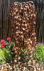 A decorative outdoor water feature with a copper-toned leaf-shaped metal cascade mounted in front of a dark bamboo screen, surrounded by green plants and colorful flowers on a bed of small pebbles.