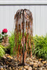 A copper-toned metal garden sculpture shaped like a weeping tree, standing in a bed of smooth river rocks with green plants and a pink flower in a garden setting.
