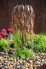 A metal weeping tree garden sculpture with a copper-toned leafy canopy stands in a landscaped garden bed with green ornamental grass, a dark brown wooden fence, and wet multicolored pebbles.
